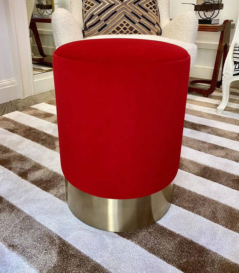 Un pouf rivestito in velluto rosso sulla base dorata, davanti a una poltrona con un cuscino grafico